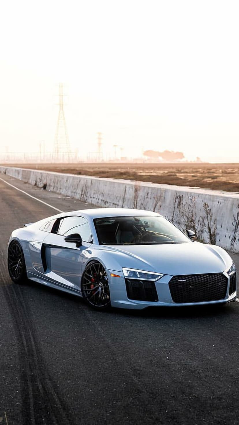Silver Audi R8 sports car parked on a paved road with tire marks against a hazy sunset and industrial horizon.