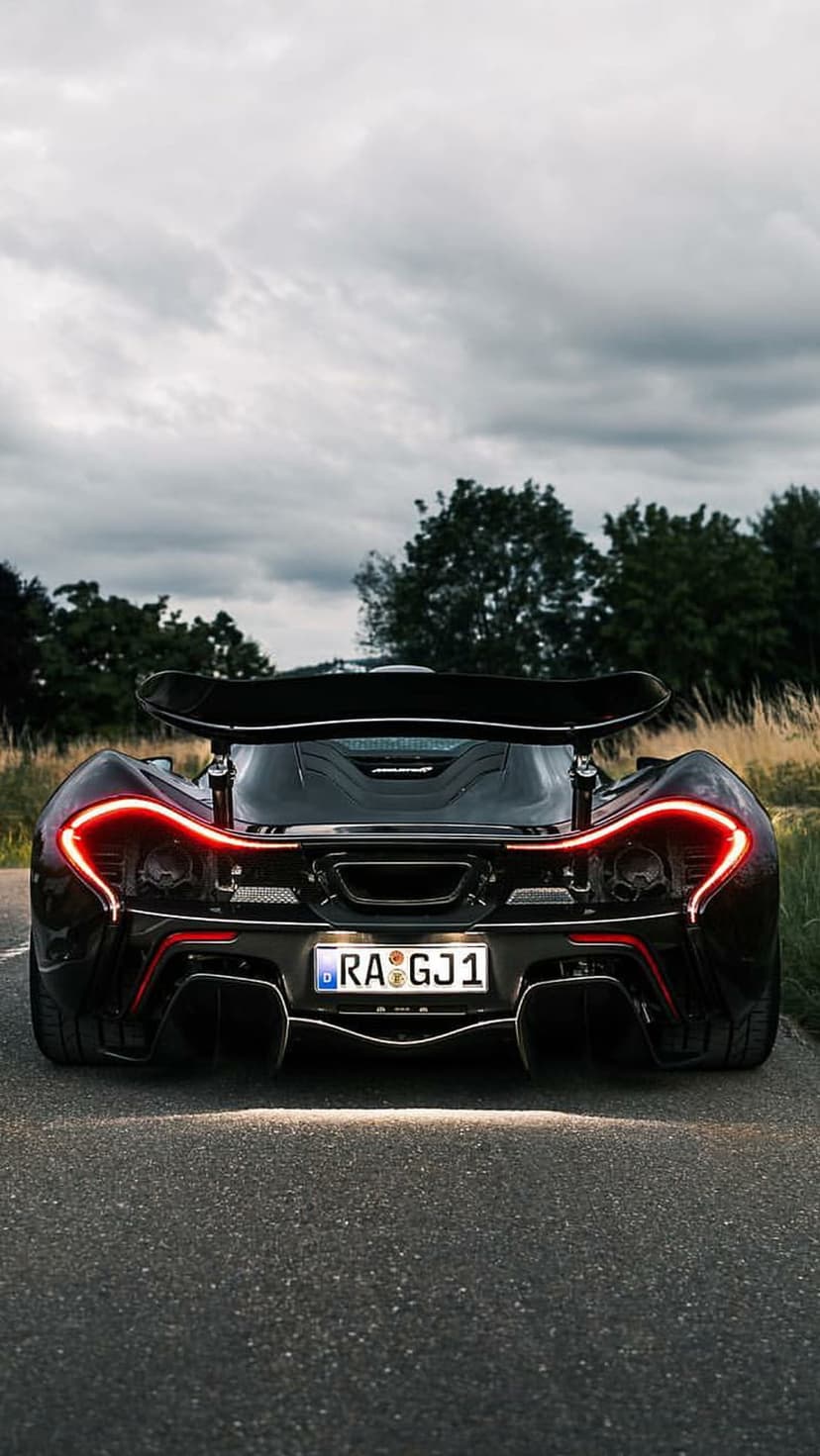 McLaren P1 hypercar rear view with circular taillights and a large wing on a paved road under a cloudy sky.