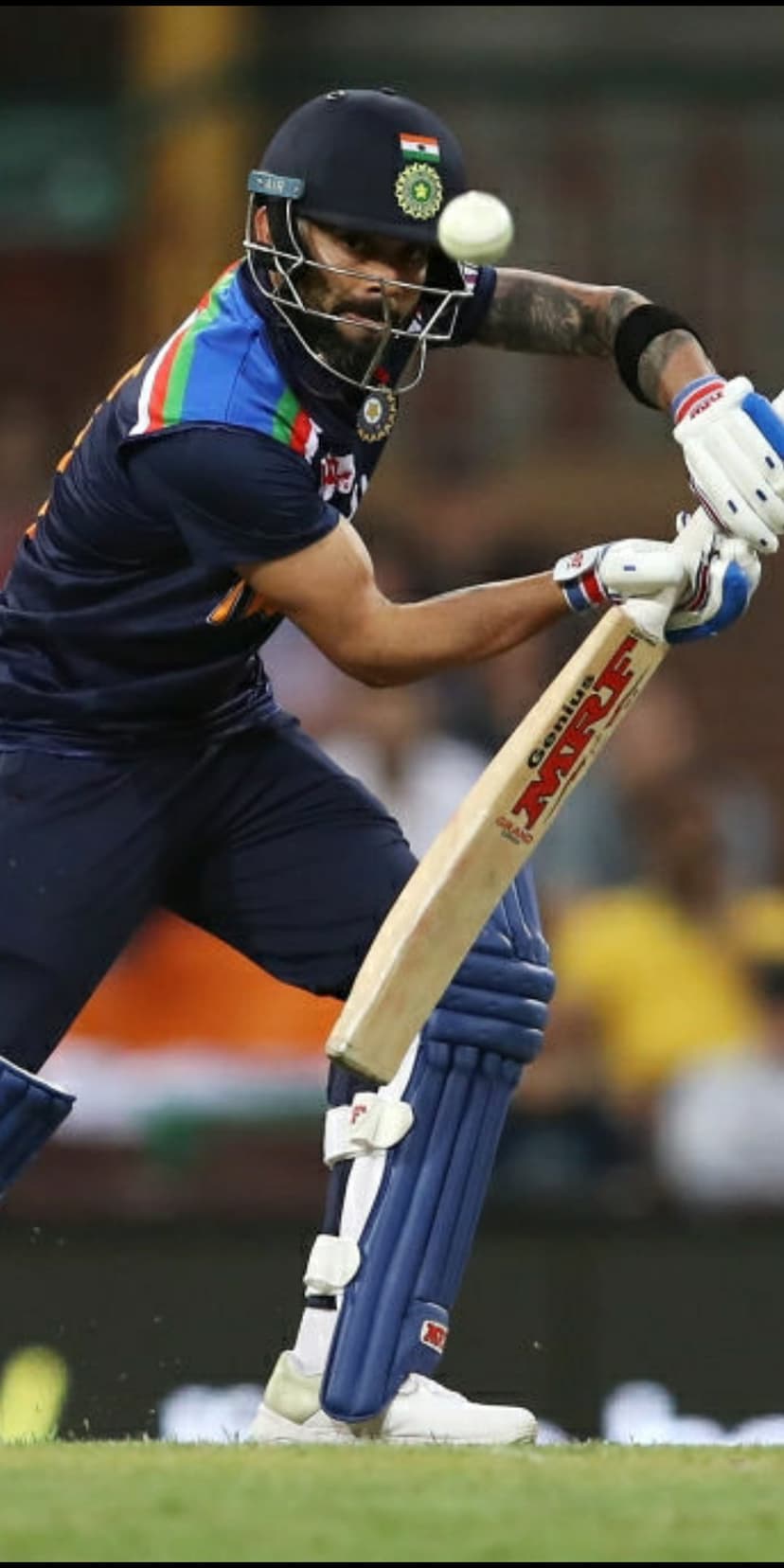 Virat Kohli follows through on a batting swing while wearing a blue India jersey and protective cricket equipment.