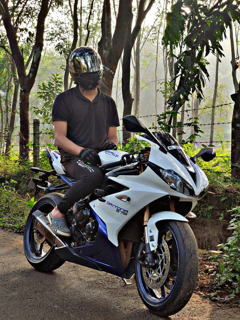 Motorcyclist in black gear sits on a blue sport bike on a forest road with sunlight filtering through trees.