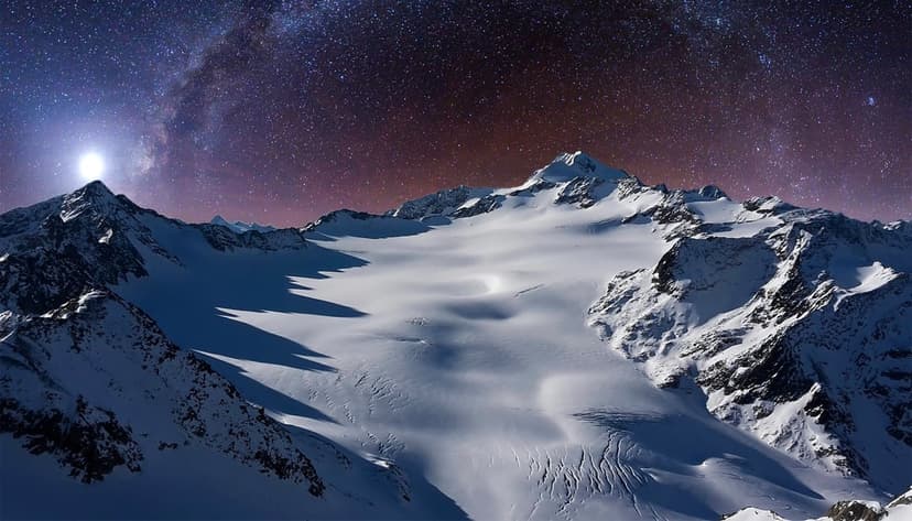 Snow-dusted mountain peaks and a sprawling glacier sit under a brilliant Milky Way galaxy in a dark night sky.