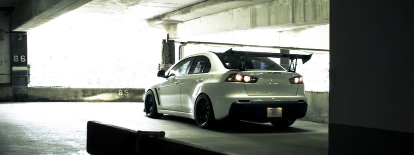 White Mitsubishi Lancer Evo X with a large rear wing parked in a concrete garage with bright side lighting.
