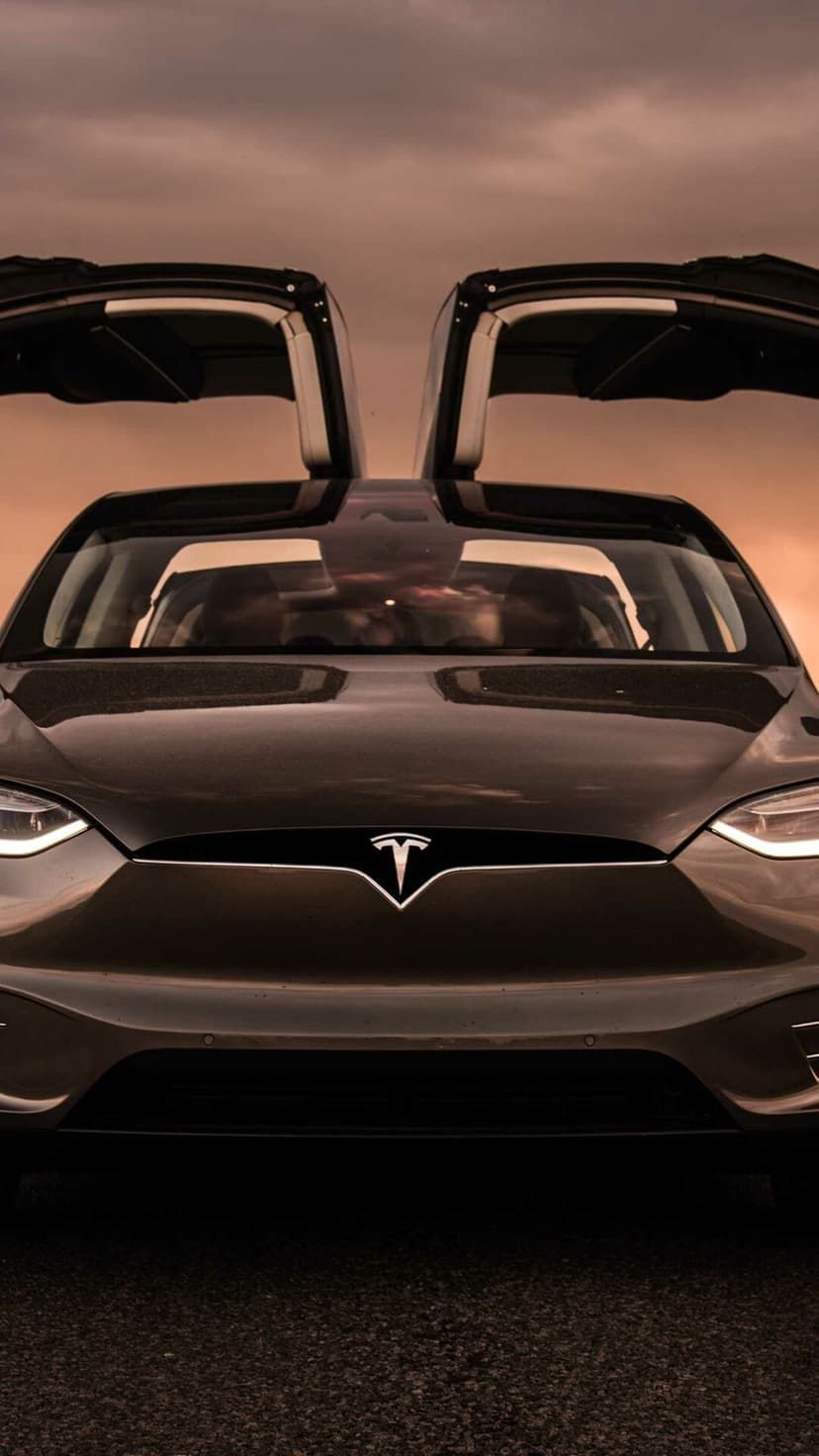 Front view of a grey Tesla Model X with open falcon doors against an orange sunset sky with glowing headlights.
