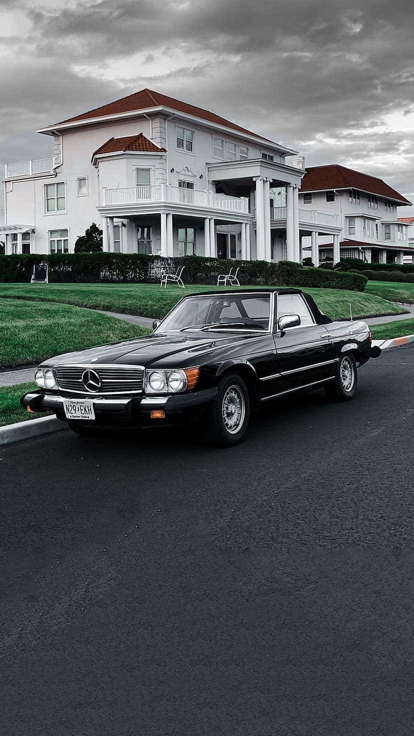 Black Mercedes SL convertible parked on a driveway in front of a white mansion with a red tile roof and lawn.