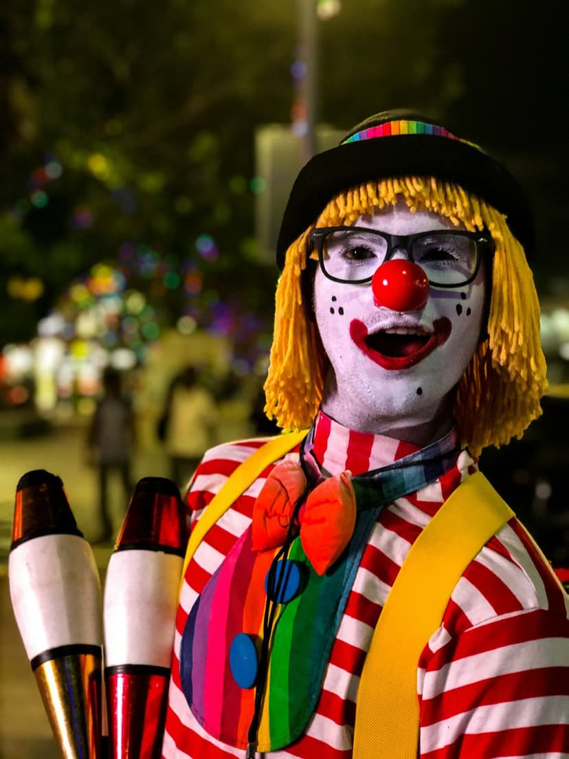 Juggling clown with a yellow wig, red nose, and rainbow jabot holds pins against a dark carnival background.