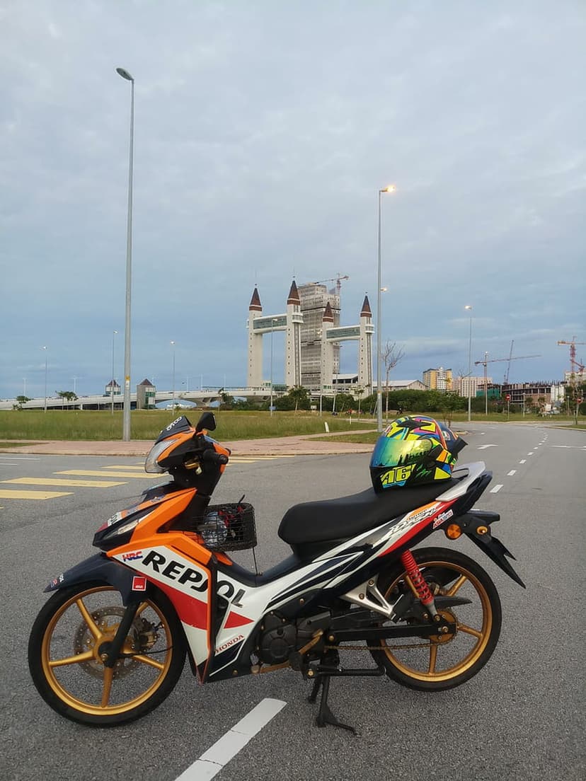 Orange motorcycle with gold rims parked on a road near a futuristic skyscraper under a heavy cloudy sky.