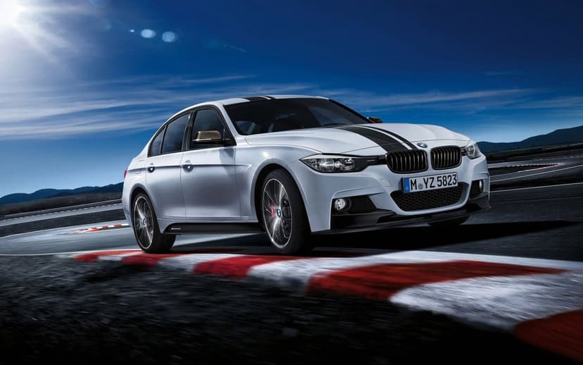 White BMW sedan with black racing stripes driving fast on a racetrack with red curbing under a sunny blue sky.