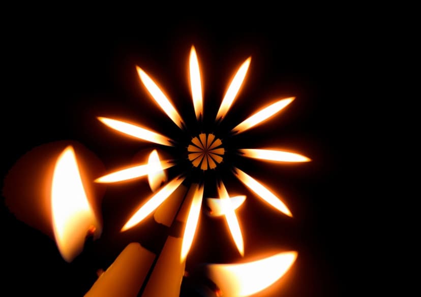 Circular arrangement of orange candle flames forming a kaleidoscopic flower pattern against a black background.