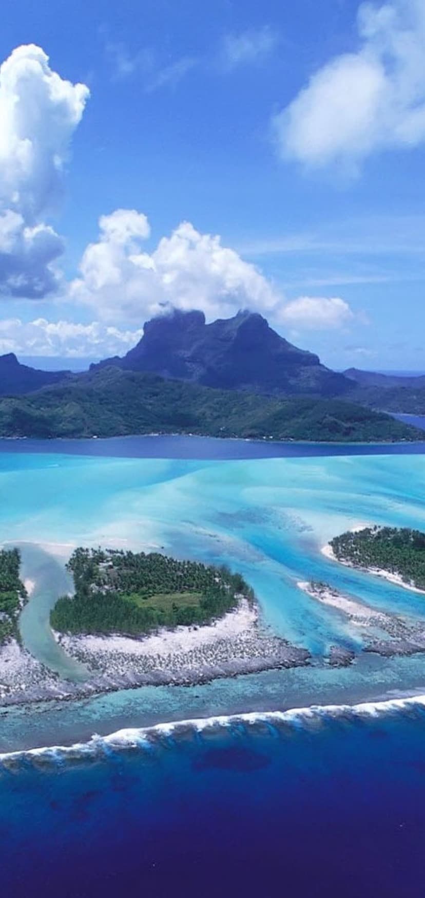 Mount Otemanu rises above a turquoise lagoon with coral reefs and green islets under a blue sky with white clouds.