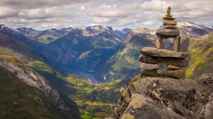 Stone cairn stands on a cliff edge overlooking a deep green mountain valley with a winding fjord under cloudy skies.