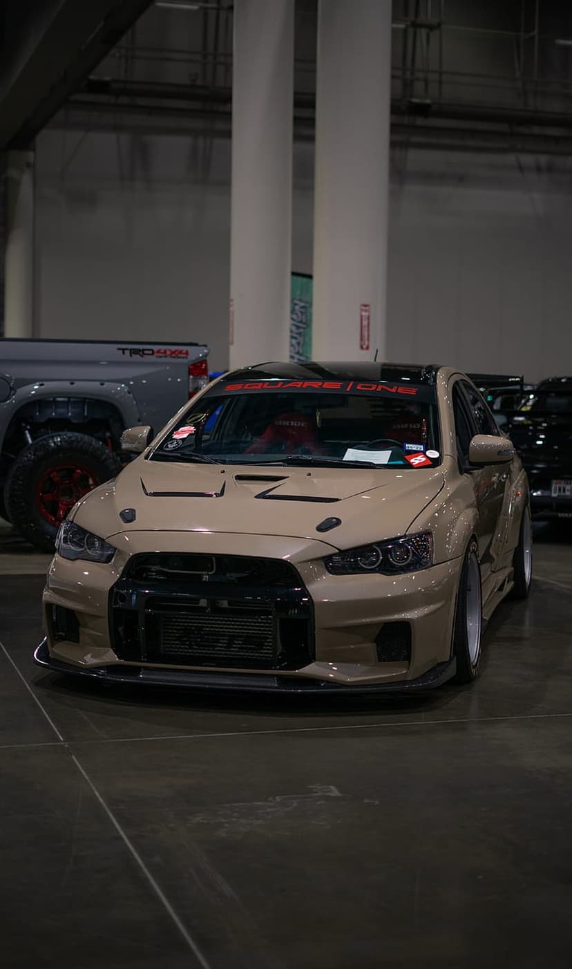 Mitsubishi Lancer Evolution X with a tan widebody kit, large intercooler, and red racing seats at a car show.