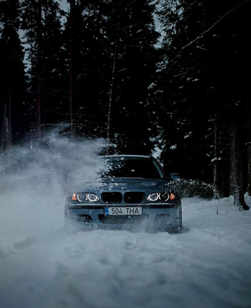 BMW sedan drifting on a snowy forest road at night with glowing headlights and high-speed powder spray.