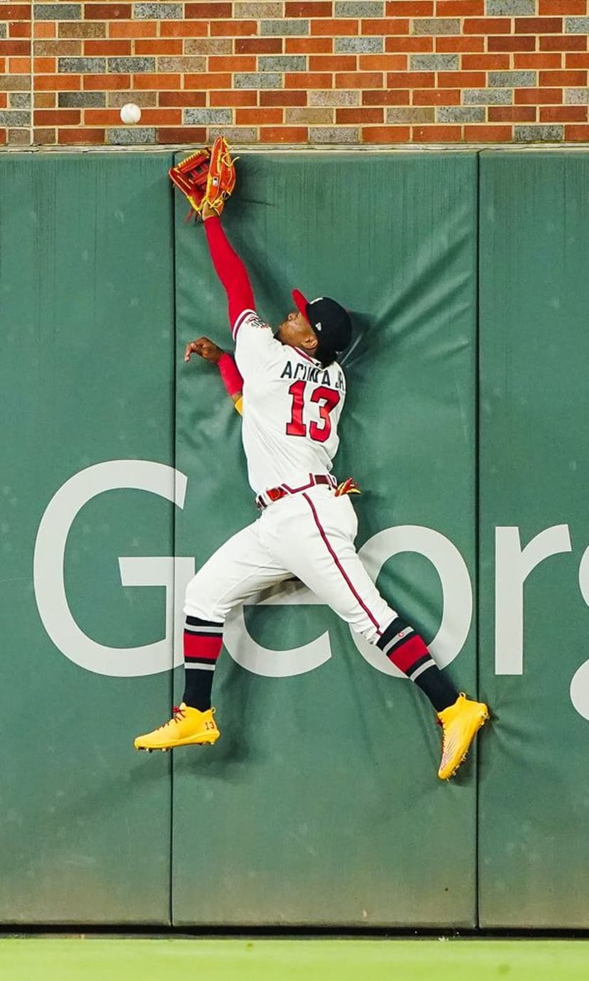 Ronald Acuña Jr. jumps to catch a baseball against a green padded fence in a vertical action sports shot.