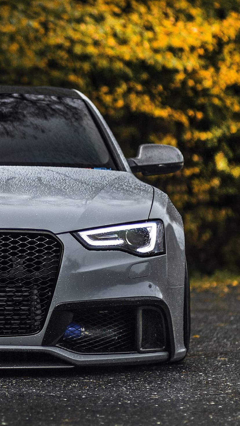 Audi RS5 convertible with glowing LED headlights on a wet road surrounded by blurred yellow autumn foliage.