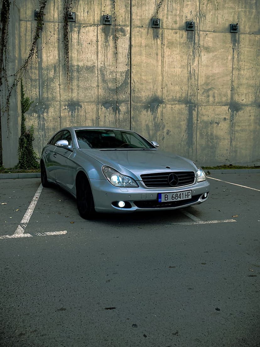 Silver Mercedes CLS sedan with glowing headlights parked before a concrete wall and ivy on an asphalt lot.