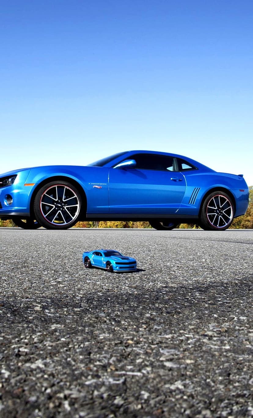 Blue Chevrolet Camaro and a small toy car version placed on an asphalt road under a clear sky with trees.
