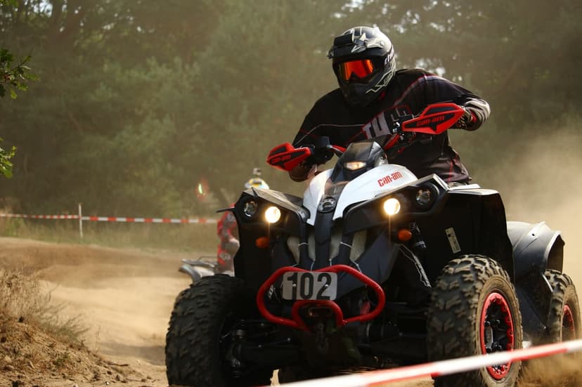 ATV racer wearing a helmet and goggles drives a quad bike through a thick cloud of dust on a dirt racing track.