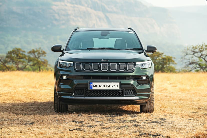 Front view of a dark green Jeep Compass SUV parked in a dry grassy field with hazy mountains in the background.