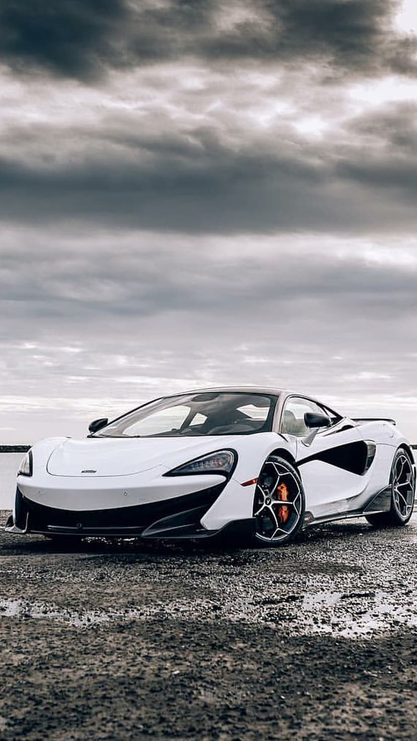 White McLaren sports car with orange calipers parked on a wet rocky shore beside a lake under heavy grey clouds.