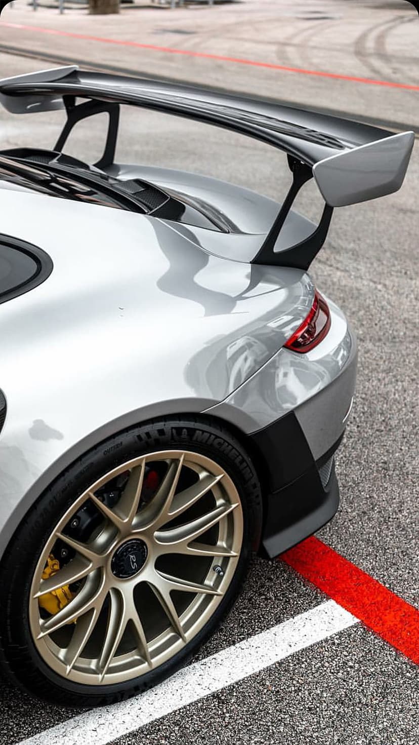 Porsche 911 GT3 RS rear view with silver paint, massive wing, gold wheels, and yellow calipers on a race track.