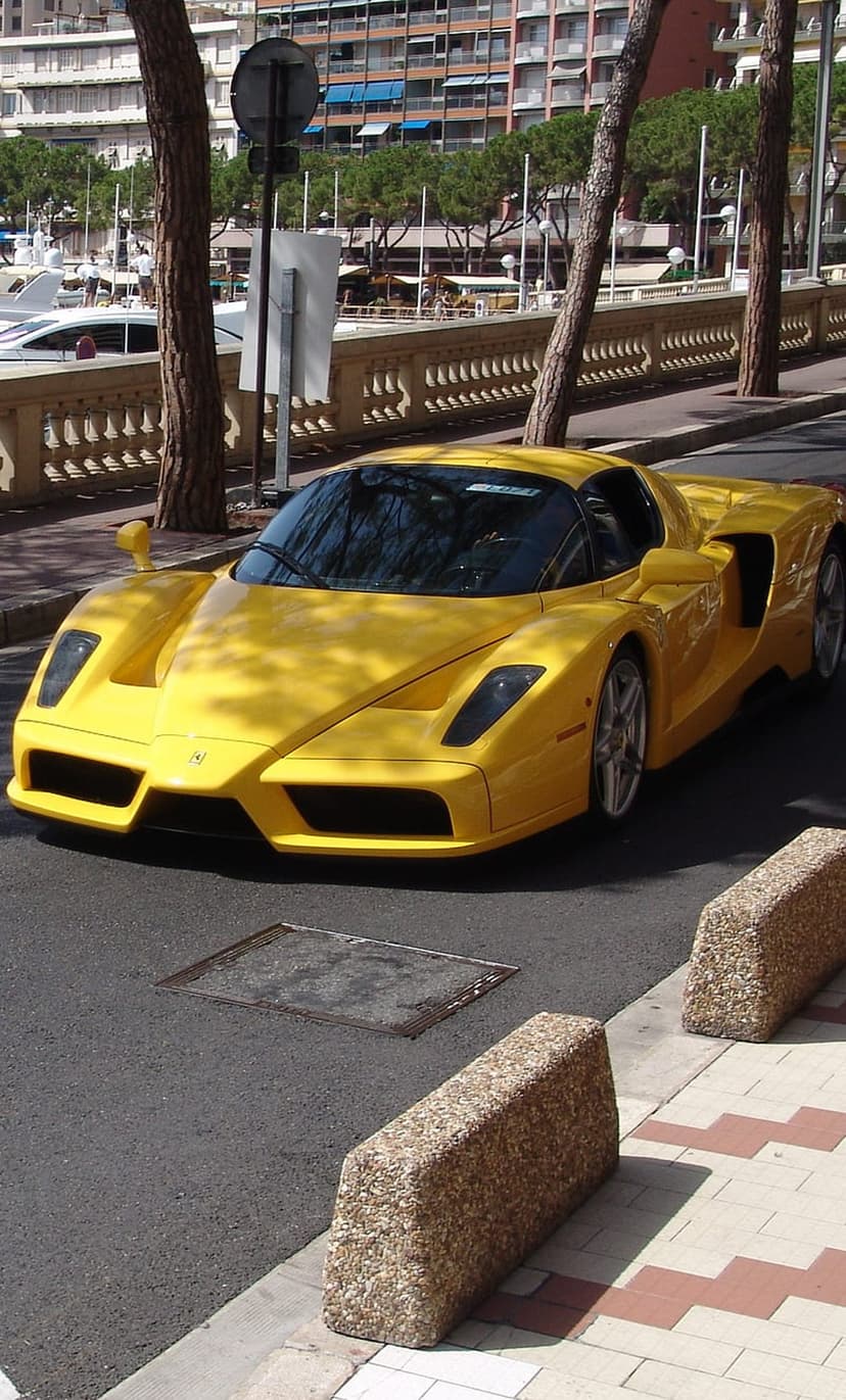 Yellow Ferrari Enzo driving on a Monaco street with palm trees and a marina filled with yachts in the background.