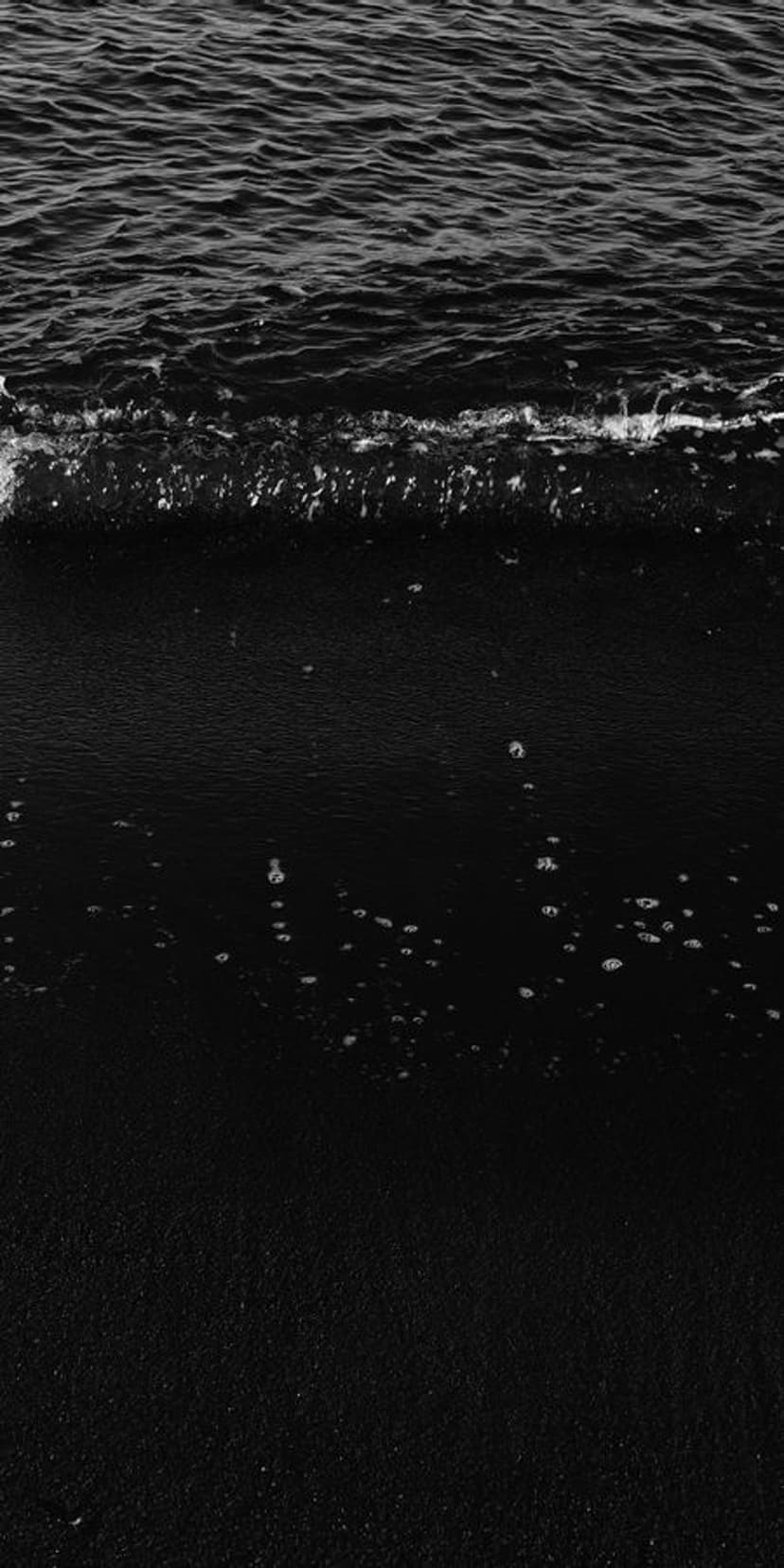 Frothy white ocean waves break against a dark volcanic sand beach with visible ripples in the receding water.