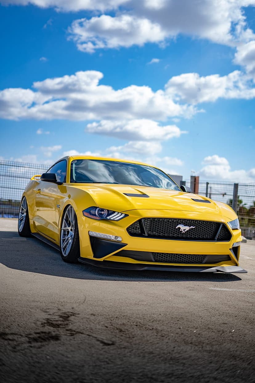 Yellow Ford Mustang convertible with its top down parked on asphalt under a sunny blue sky with white clouds.