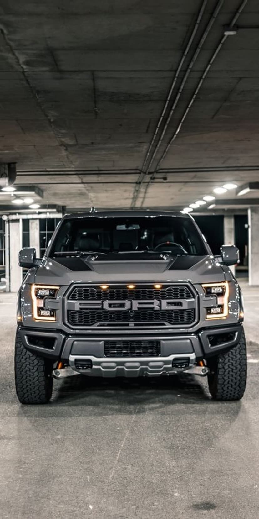Dark grey Ford F-150 Raptor truck with bold front lettering parked in a shadowy concrete parking garage.
