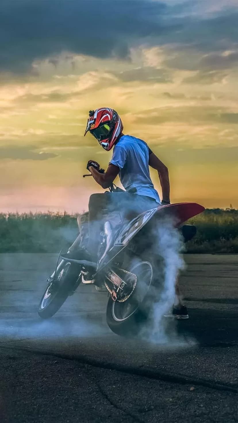 Biker in a blue shirt lifts the front wheel of a motorcycle while creating a smoke trail during a bright sunset.