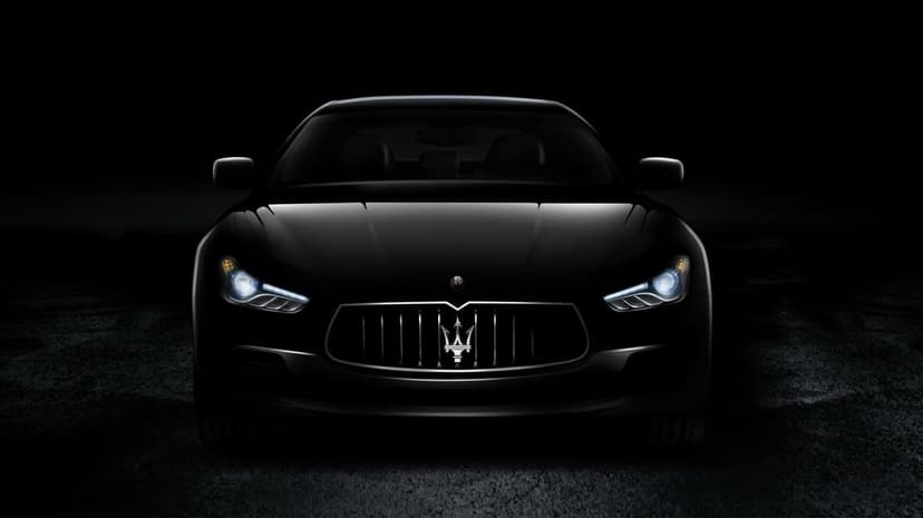 Front view of a black Maserati Ghibli with glowing headlights and trident logo in a dark, moody environment.