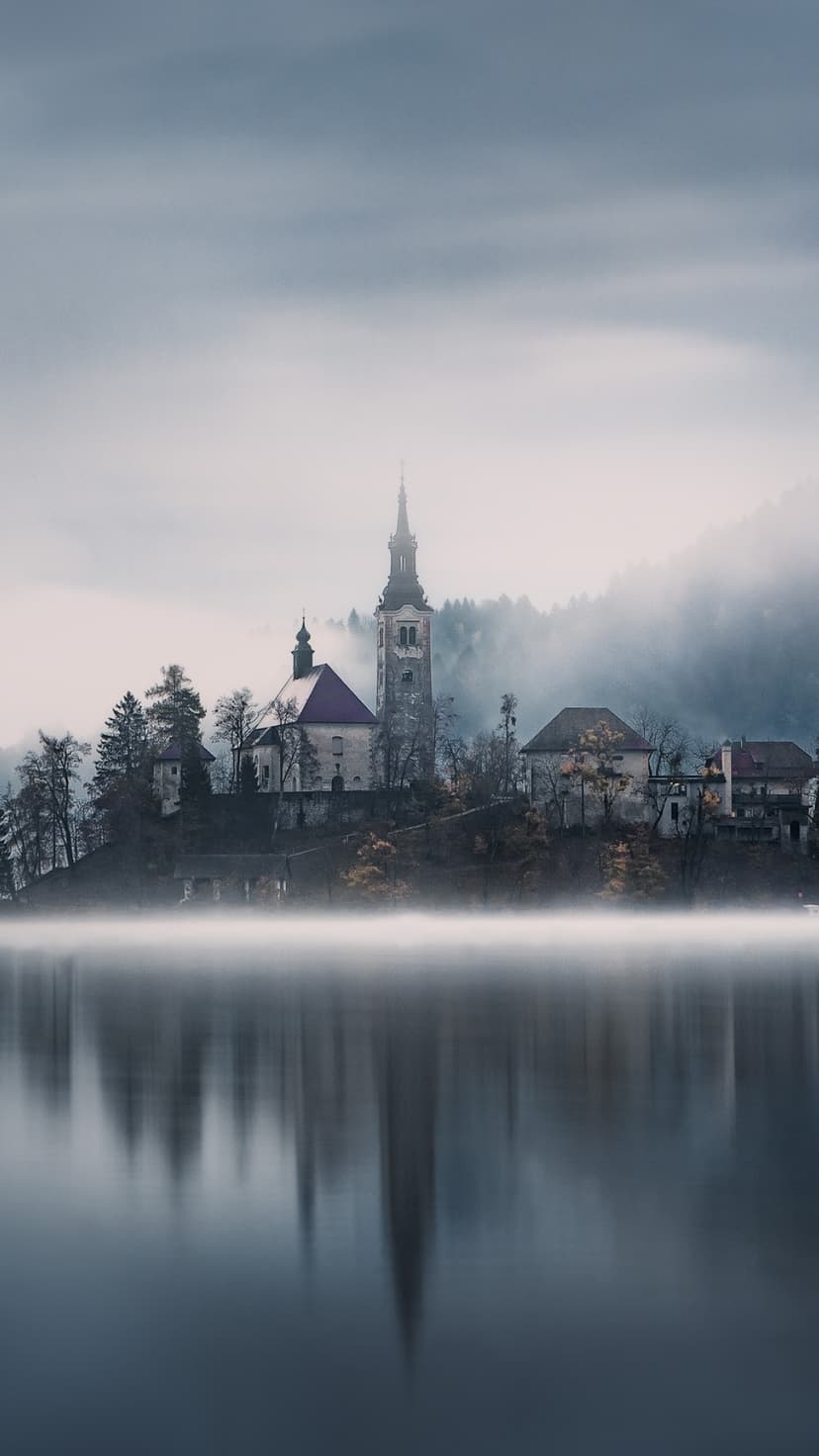 Church spire emerges from dense white fog on a small island over still lake water with mountain silhouettes.