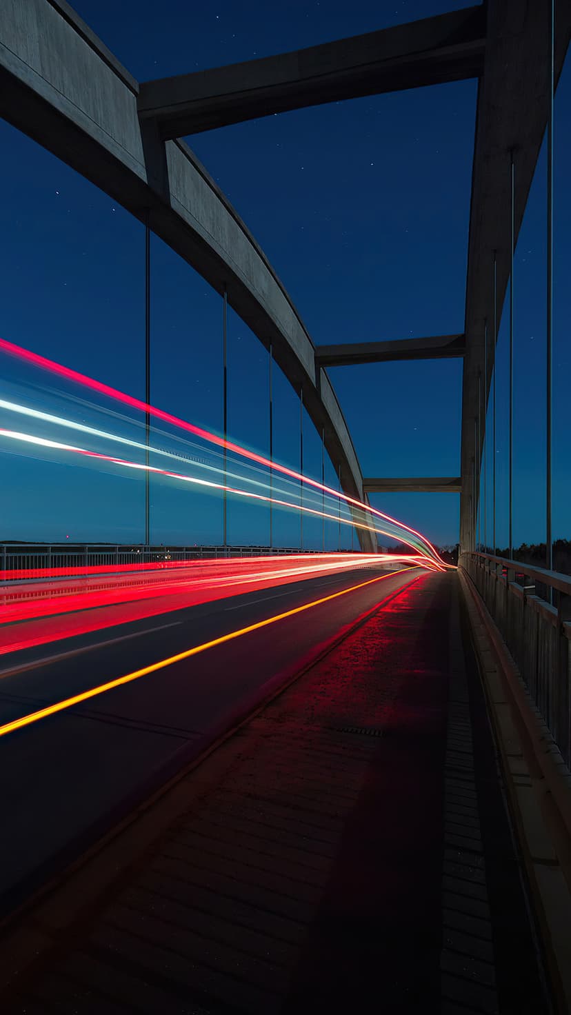 Concrete arch bridge with glowing red and white vehicle light trails under a dark blue twilight sky at night.