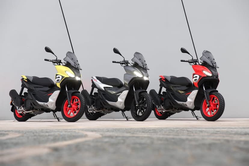 Three Aprilia SR GT scooters in yellow, silver, and red with headlights on against a grey wall background.