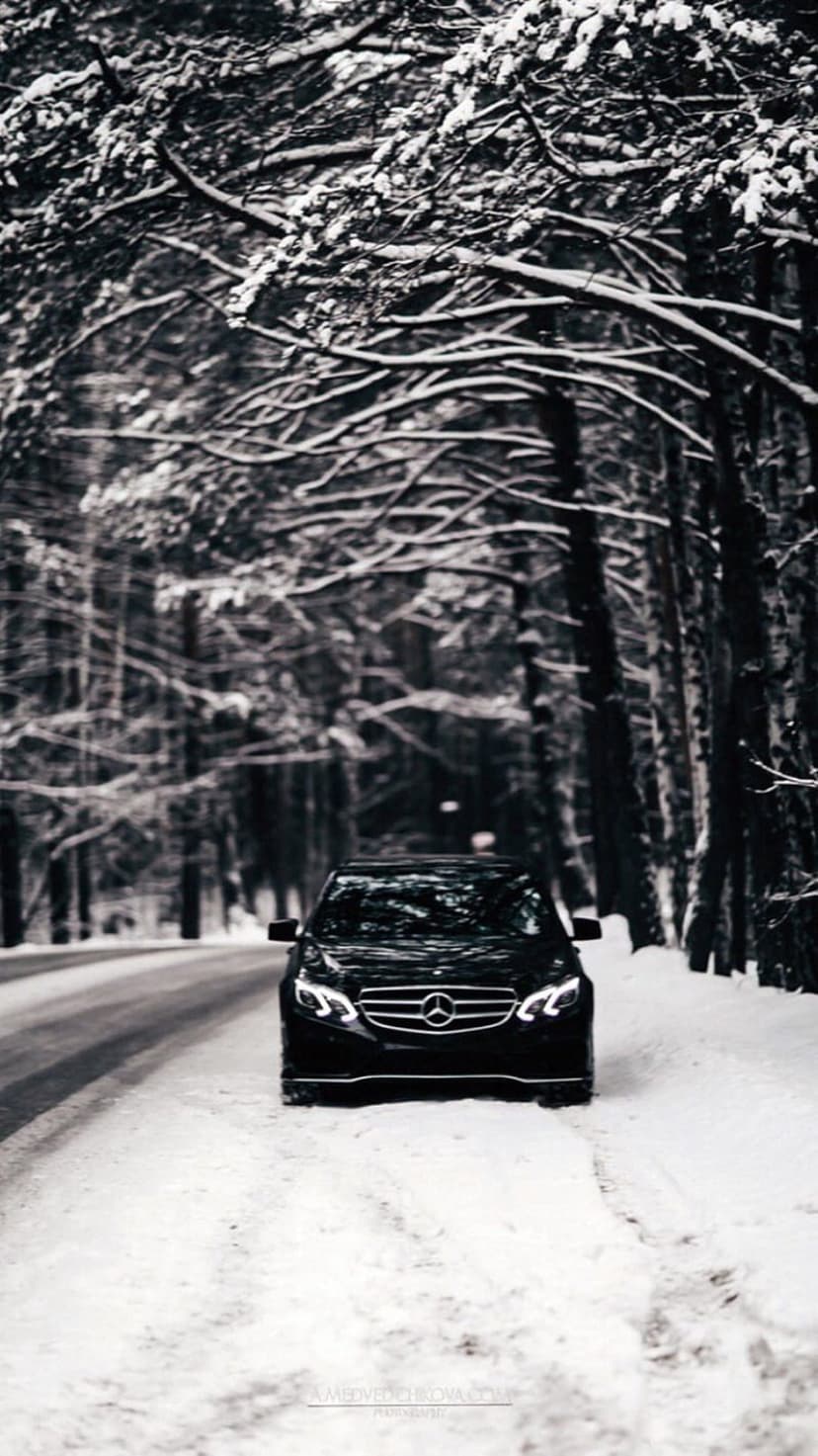 Black Mercedes-Benz sedan cruises through a snow-covered forest with bright headlights on a frozen road.