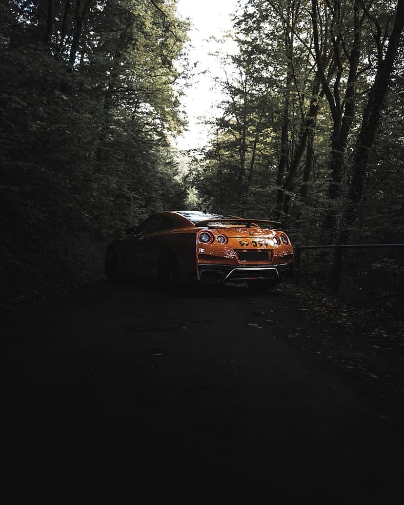 Orange Nissan GT-R R35 parked on a dirt trail under towering trees with sunlight filtering through the leaves.