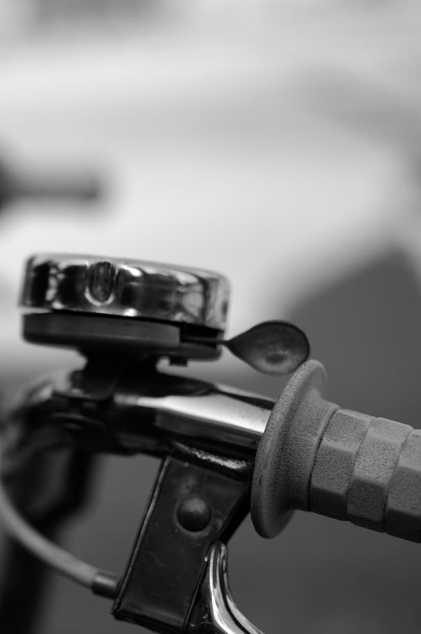 Polished chrome bicycle bell with a mechanical lever mounted on a textured rubber handlebar in monochrome.