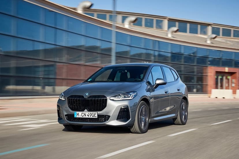 Grey BMW 2 Series Active Tourer driving on a sunlit road past a modern glass building with motion blur.
