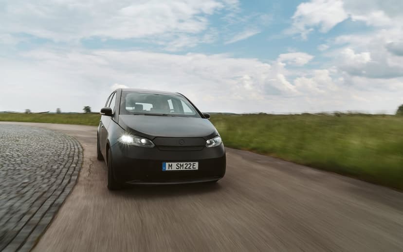 Matte black electric car with glowing headlights driving on a blurred rural road under a bright cloudy sky.