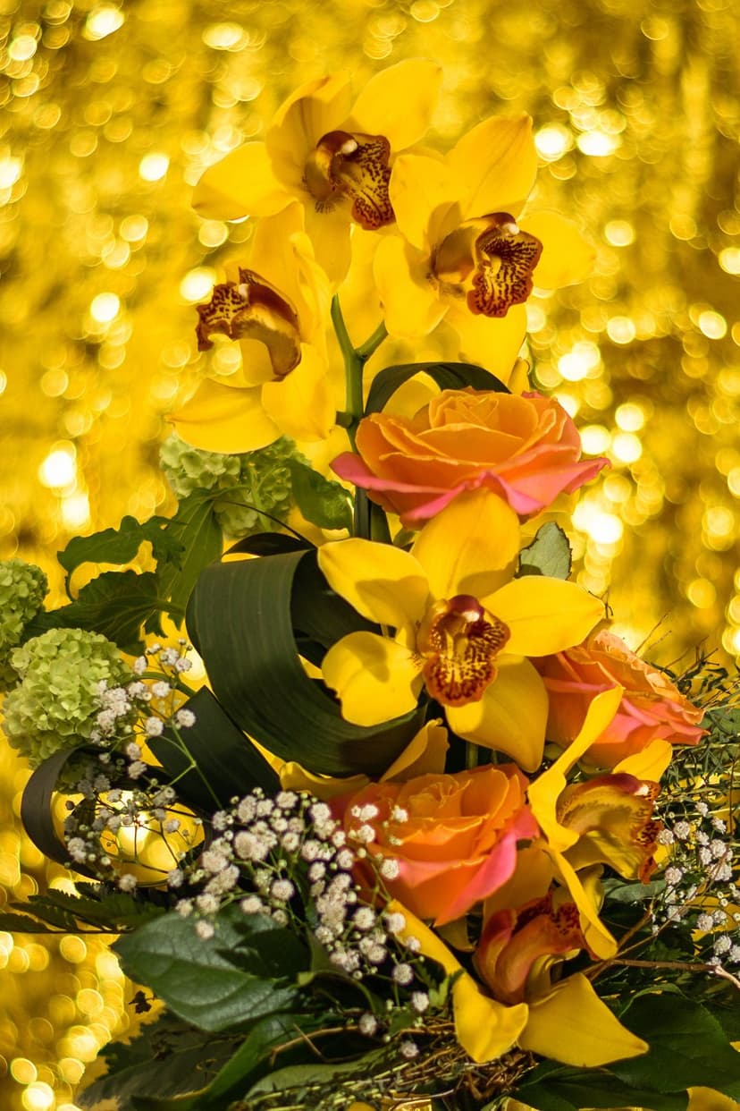 Yellow orchids and orange roses in a dense bouquet set against a soft, shimmering golden bokeh background.