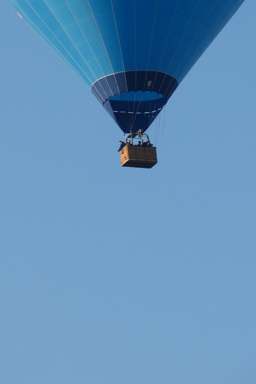 Blue hot air balloon with a wicker basket floats upward through a vast, cloudless azure sky on a clear day.