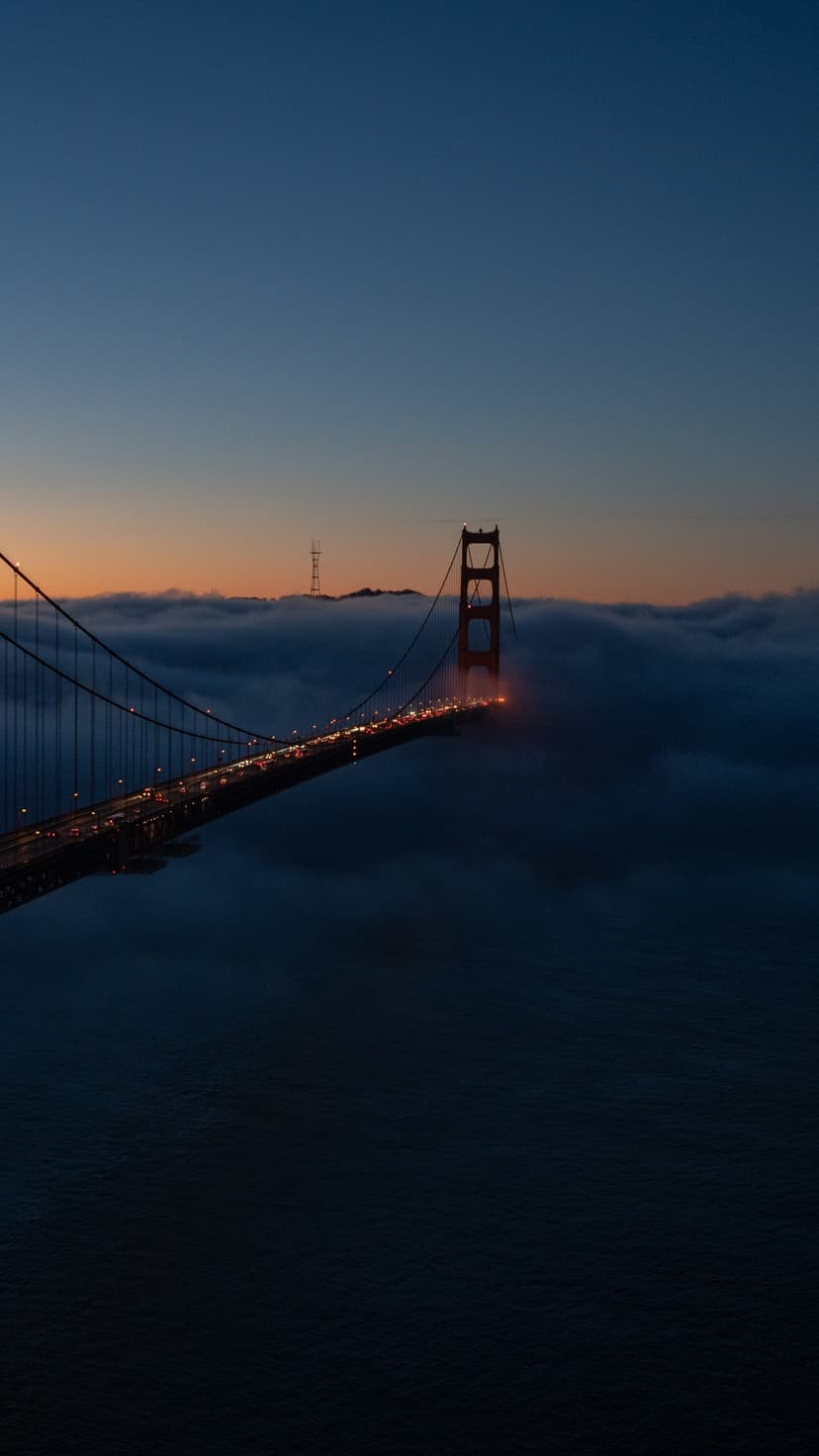 Golden Gate Bridge towers rise from a thick layer of fog at twilight under a gradient orange and blue sky.