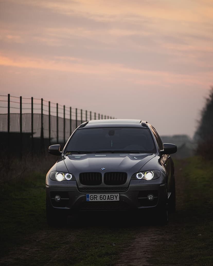 BMW X6 SUV with illuminated headlights parked on a dirt road against a pastel sunset sky and rural trees.
