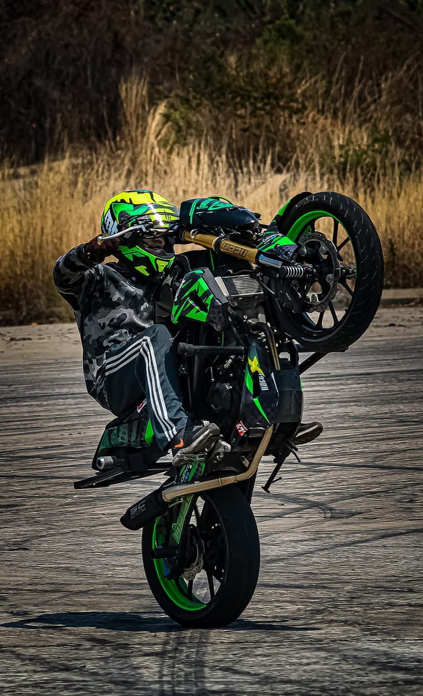 Motorcyclist in camo gear performs a wheelie on a black and green sportbike during an outdoor stunt performance.