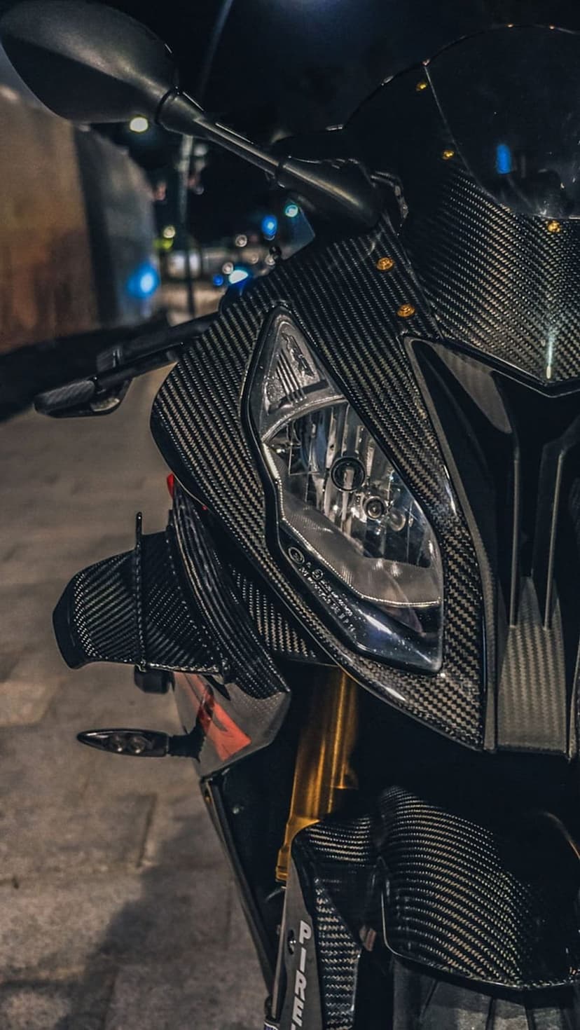 Carbon fiber motorcycle headlight housing and gold suspension forks with soft urban bokeh lights in the back.