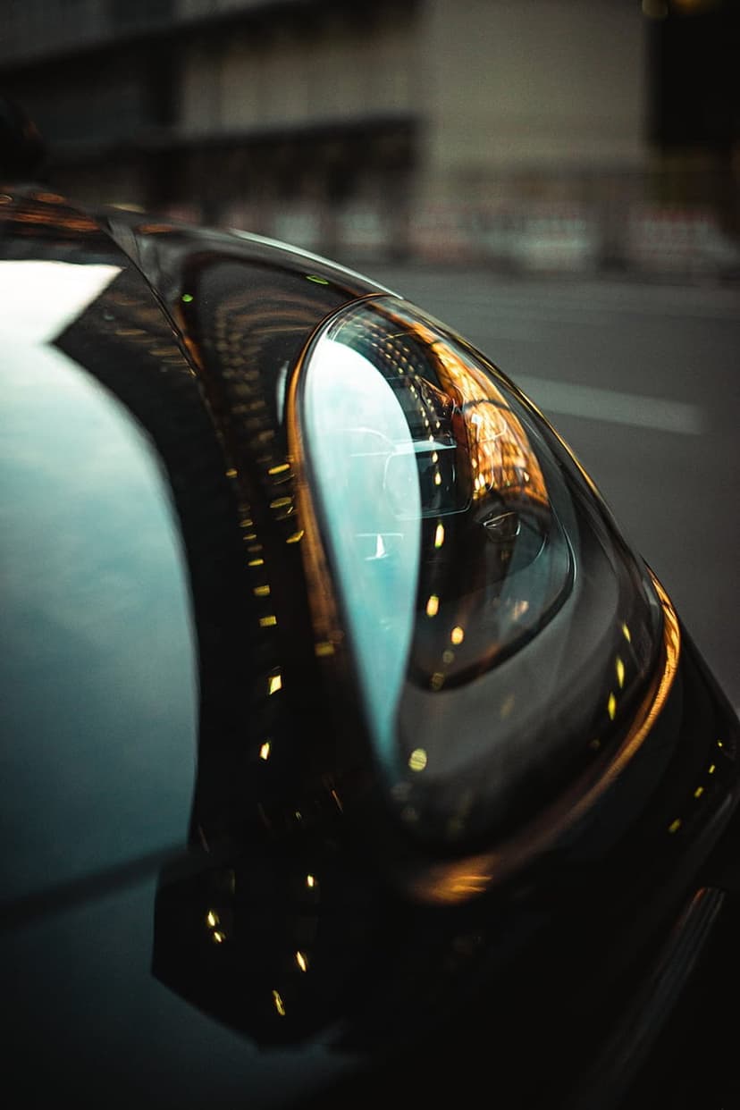 Sleek black car headlight reflecting colorful urban neon lights and bokeh against a dark metallic exterior.