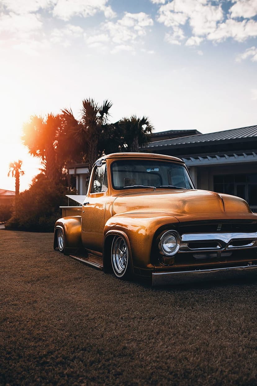 Golden vintage pickup truck with chrome wheels parked near palm trees under a warm sunset sky in an urban setting.