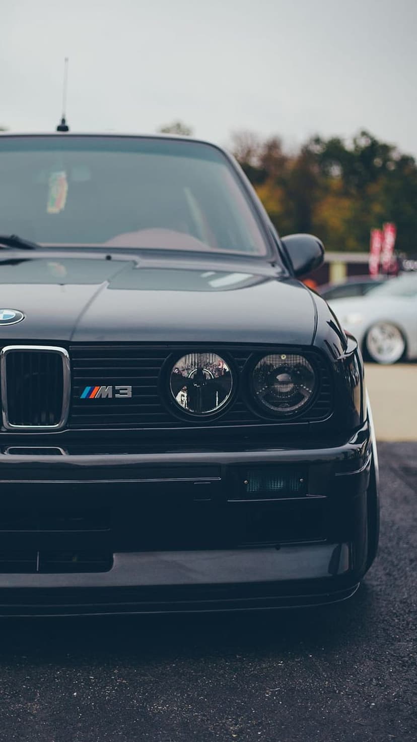 Dark gray BMW E30 M3 front grille and circular headlights with the M3 logo badge on a vintage car chassis.