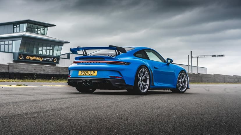 Blue Porsche 911 GT3 with a large rear wing on a racetrack under a cloudy grey sky with a control tower behind.