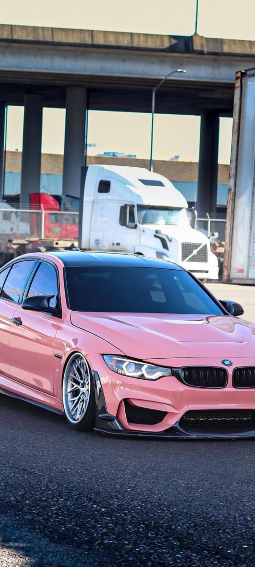 Pink BMW M4 with a black roof and custom wheels parked near semi-trucks under an industrial overpass.