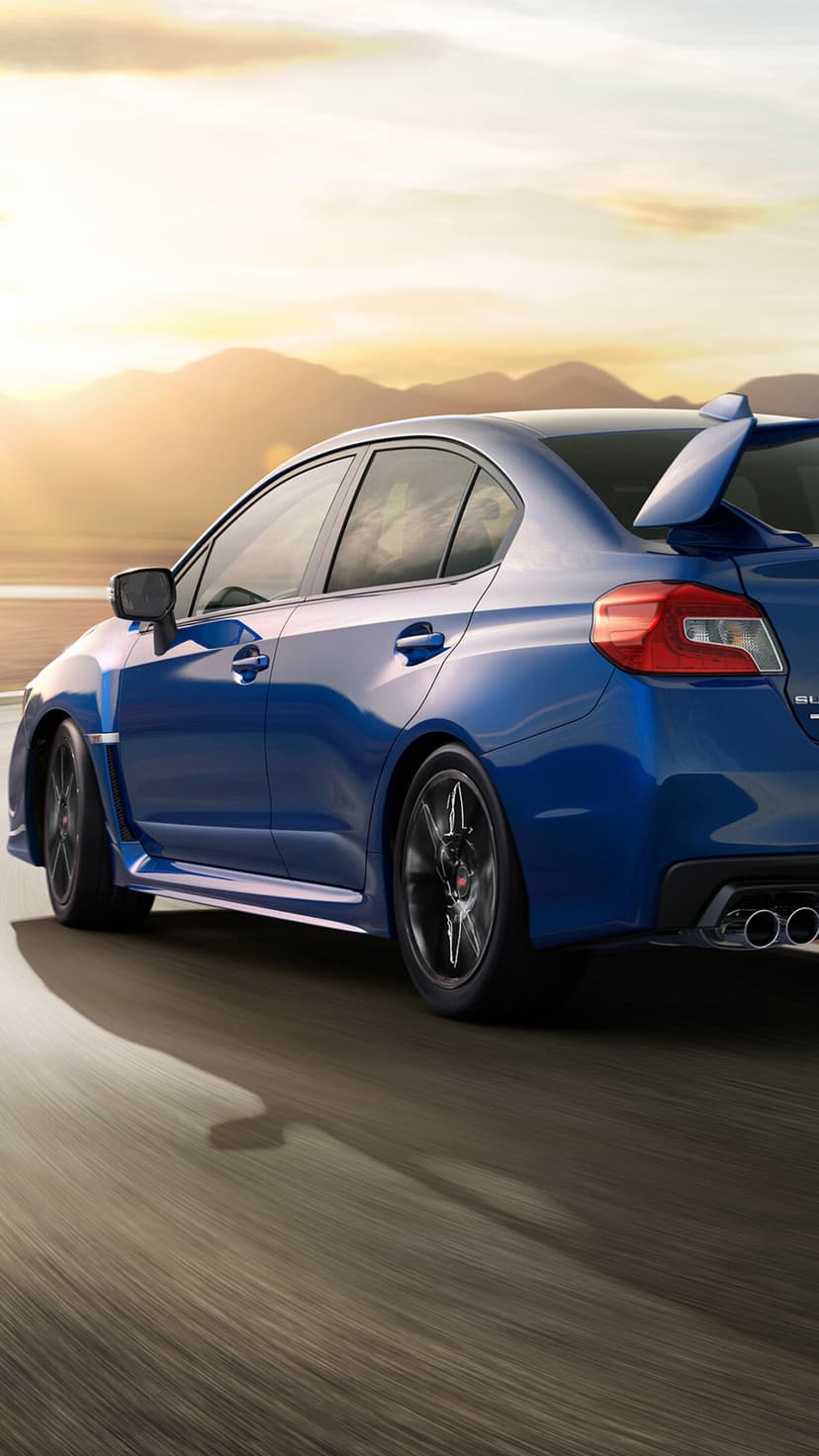 Blue sports sedan with a large rear wing driving on a curved road at sunset with mountain silhouettes.